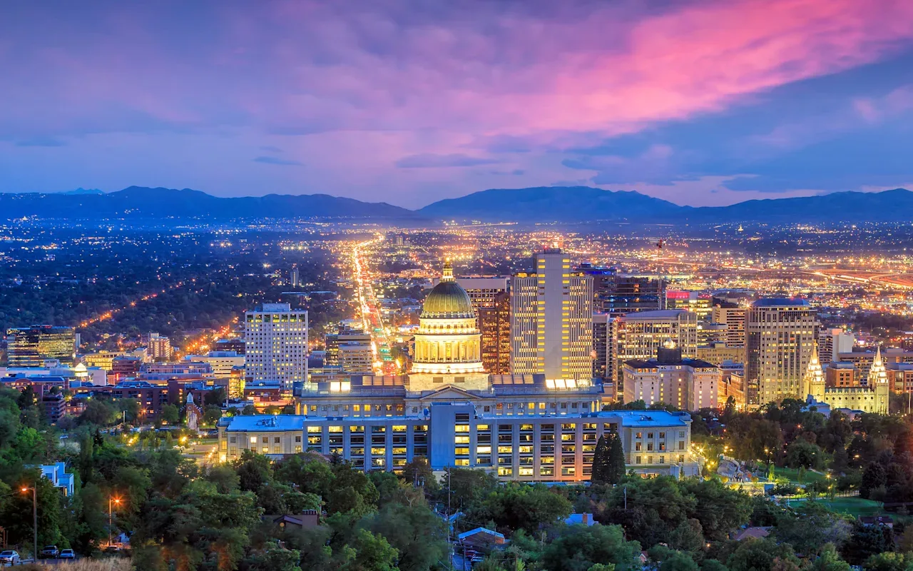 Salt Lake City at dusk from the north: the lit Capitol dome in front of downtown, the valley's lights running south, and the Wasatch range under a pink sky